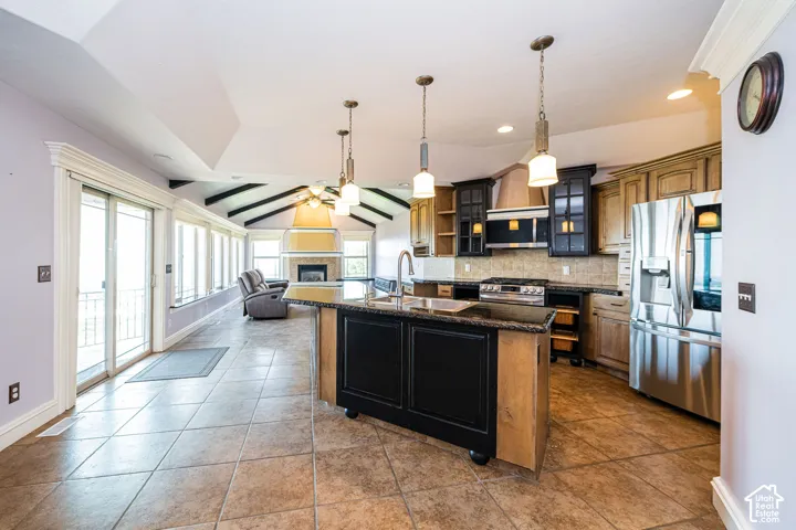 Spacious kitchen and living area featuring tile flooring, a central island with a dark base and light wood accents, stainless steel appliances, and a vaulted ceiling with exposed beams