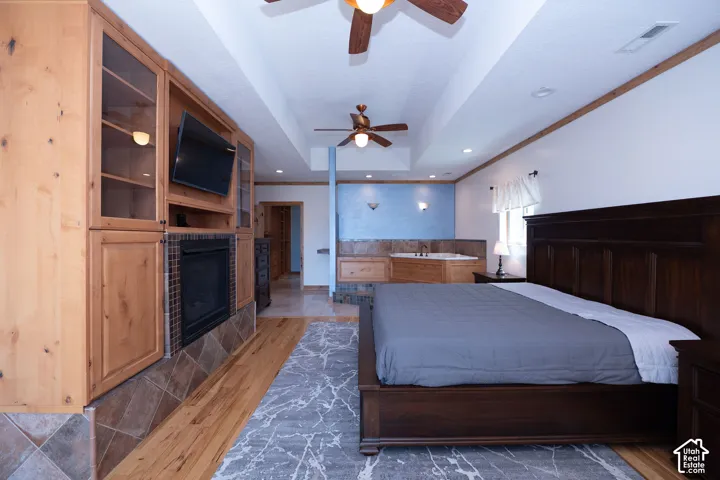 Open-concept master bedroom featuring wood-finish flooring, a built-in media center with integrated fireplace, and a tiled step-up area with an integrated jetted tub
