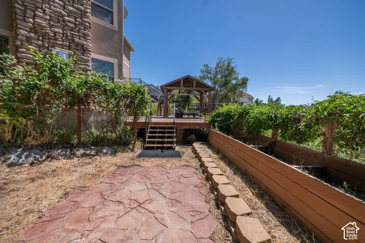 Expansive outdoor area featuring a stone-look patio, terraced landscaping with a raised planter box, and a wood deck with steps leading to a gazebo