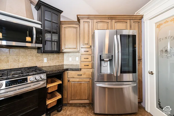 Kitchen featuring stainless steel appliances, varied wood cabinetry, a tile backsplash, dark countertops, and a frosted glass pantry door