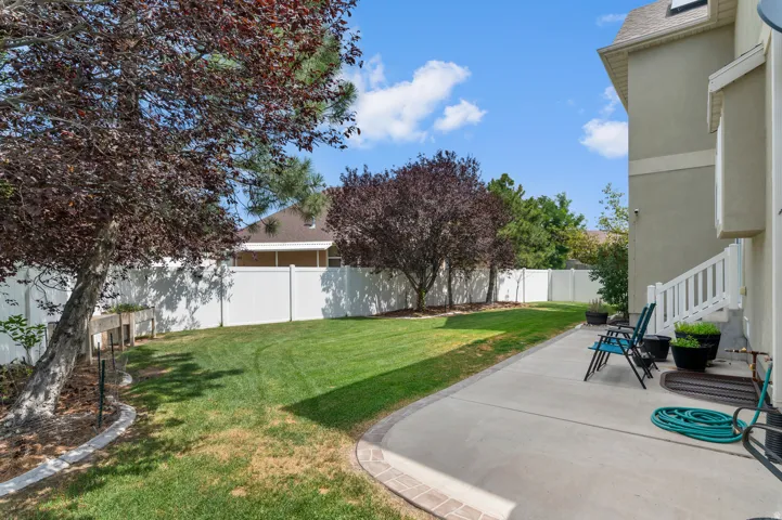 Spacious backyard featuring a concrete patio, expansive lawn, mature trees, and a white privacy fence