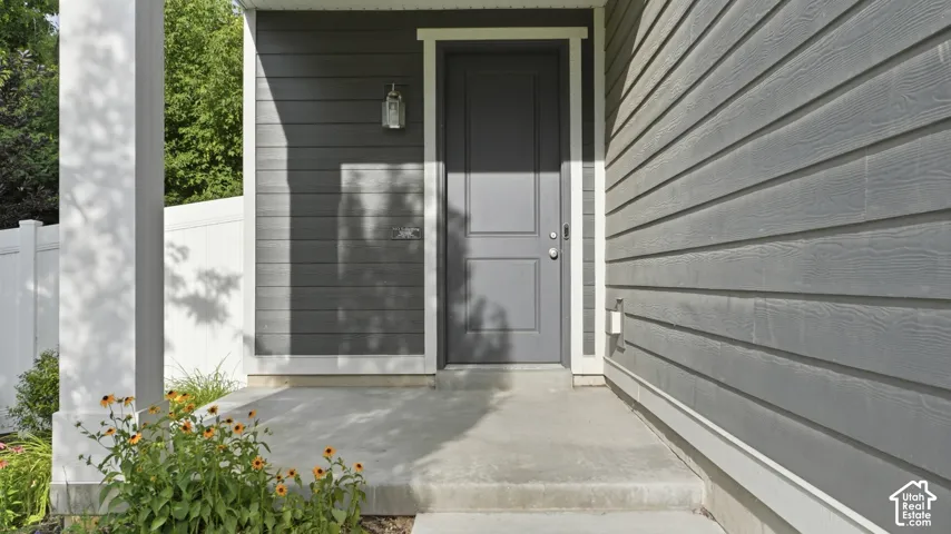 Charcoal gray front door with white trim beneath a covered porch, flanked by mature landscaping and a black wall-mount light fixture.