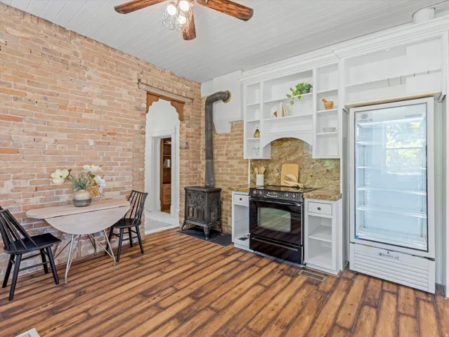 Exposed brick wall in kitchen area featuring wood-finish flooring, a wood-burning stove, and built-in shelving with an integrated range and counter space