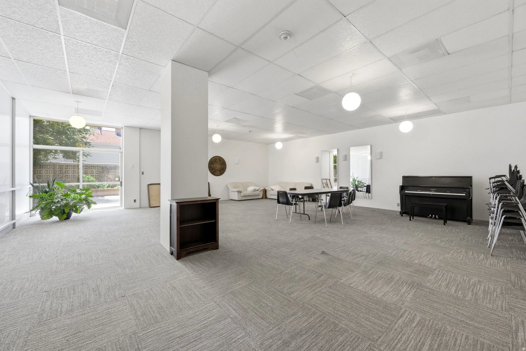 Spacious common room featuring light gray patterned carpeting, recessed ceiling lighting, and a prominent white architectural column