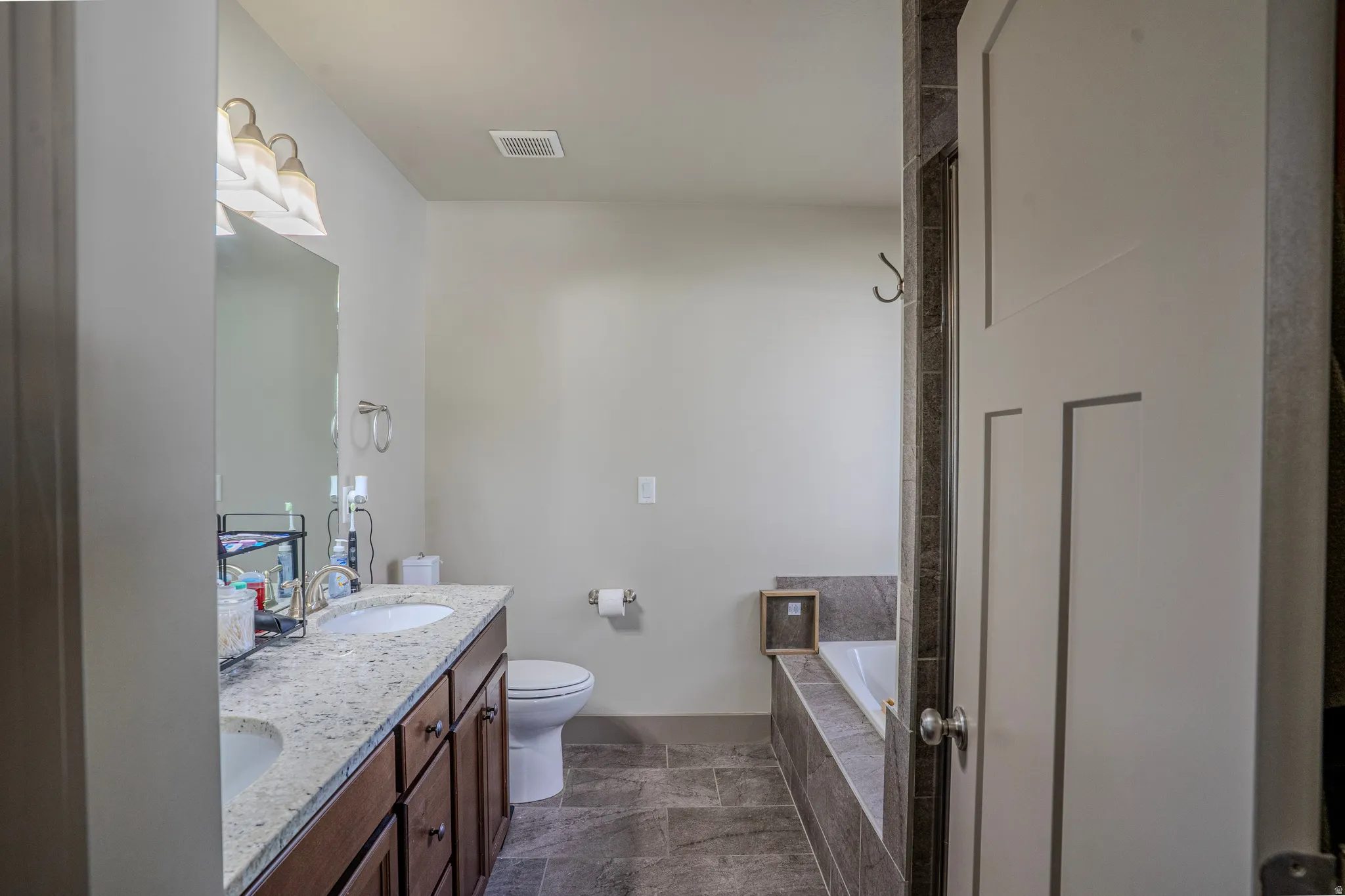 Double vanity with undermount sinks and light-toned countertop, wood-finish cabinetry, a drop-in tub with tile surround, and large format tile flooring