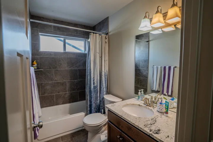Full bathroom featuring a white tub with dark gray tile surround, an undermount sink set in a light-toned countertop, a wood-finish vanity with cabinet storage, and a polished chrome faucet set