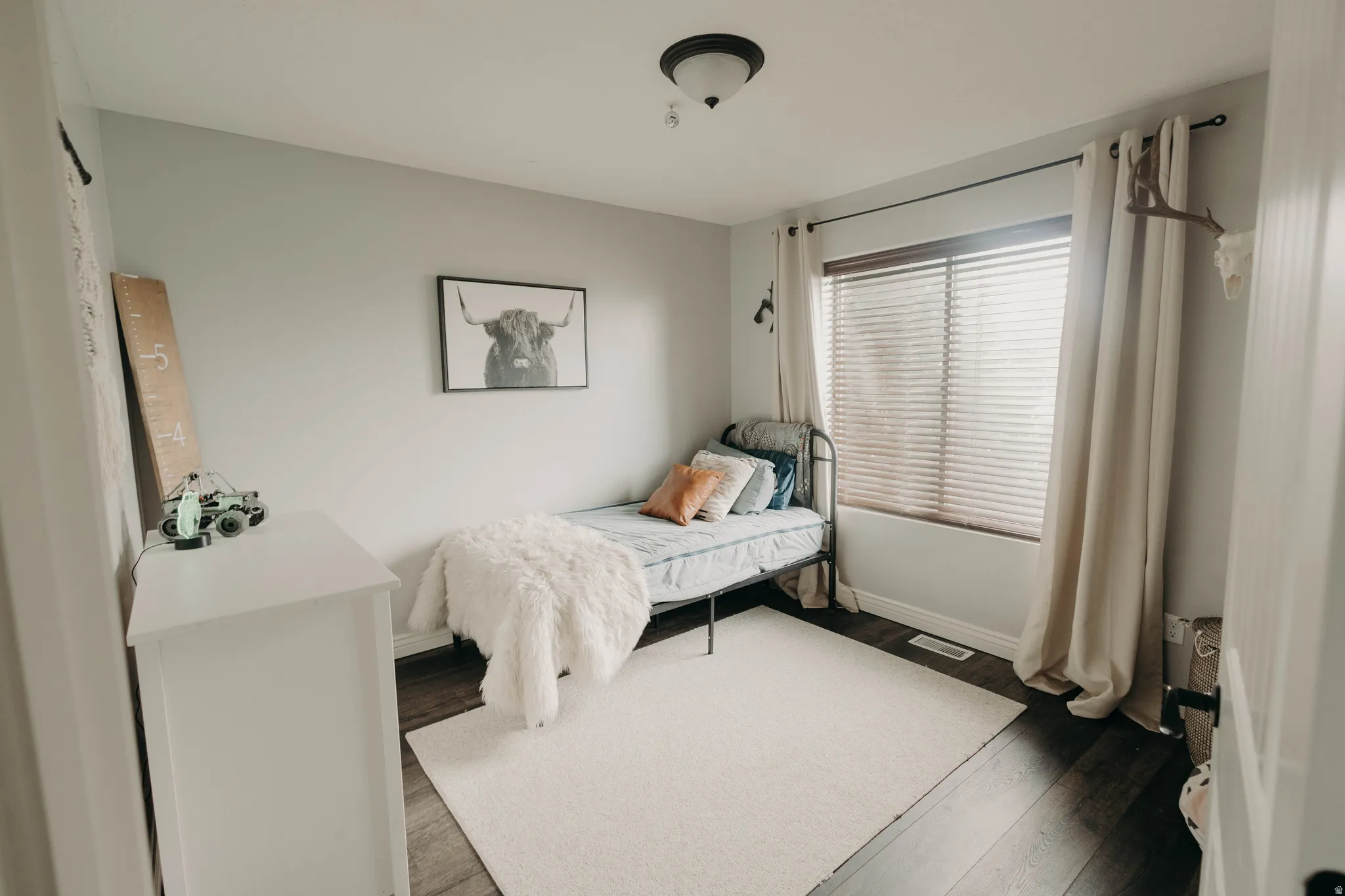 Bright interior room featuring a window with wooden blinds, hardwood-style flooring, a white area rug, and light-toned walls