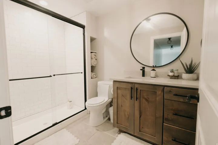 Shower with sliding glass doors and subway tile surround, wood-finish vanity with a light-toned countertop, round black-framed mirror, contemporary black plumbing fixtures, and integrated wall shelving