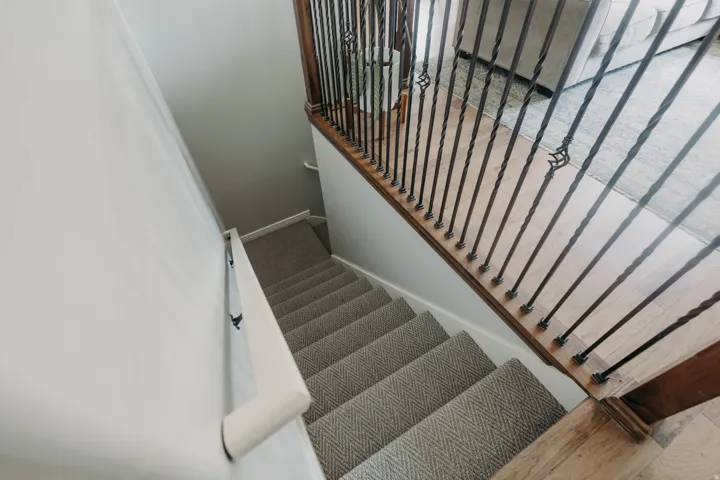 Staircase with herringbone pattern carpet, wood-finish treads, and wrought iron balusters featuring decorative accents