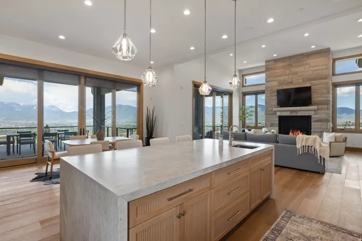 Open-concept living space featuring a large kitchen island with light-colored countertop, wood-finish flooring, a stone-clad fireplace, and expansive glass sliding doors opening to an outdoor patio with mountain scenery