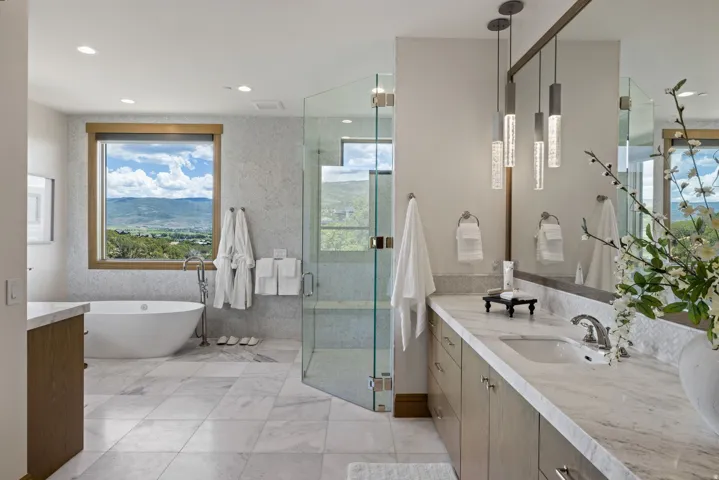 Spacious bathroom featuring a freestanding soaking tub, large window with outdoor scenery, glass-enclosed shower, dual vanity with undermount sink, and contemporary pendant lighting