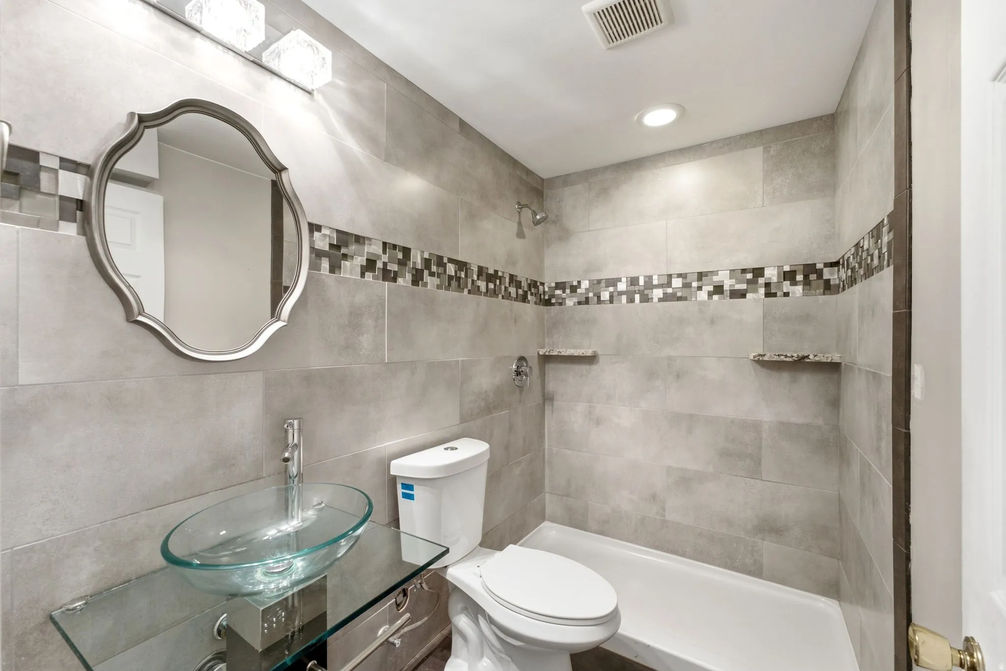 Bathroom featuring full-height tiled walls, a decorative mosaic tile band, and a glass vessel sink set on a glass vanity
