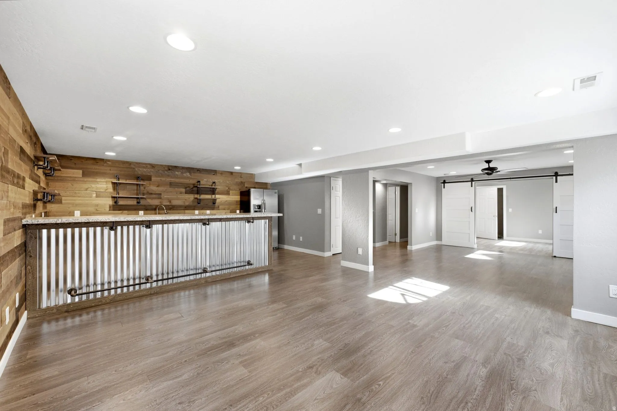 Open-concept room featuring wood-finish flooring, a corrugated metal and wood-paneled bar with a light-colored countertop, recessed lighting, and white barn doors