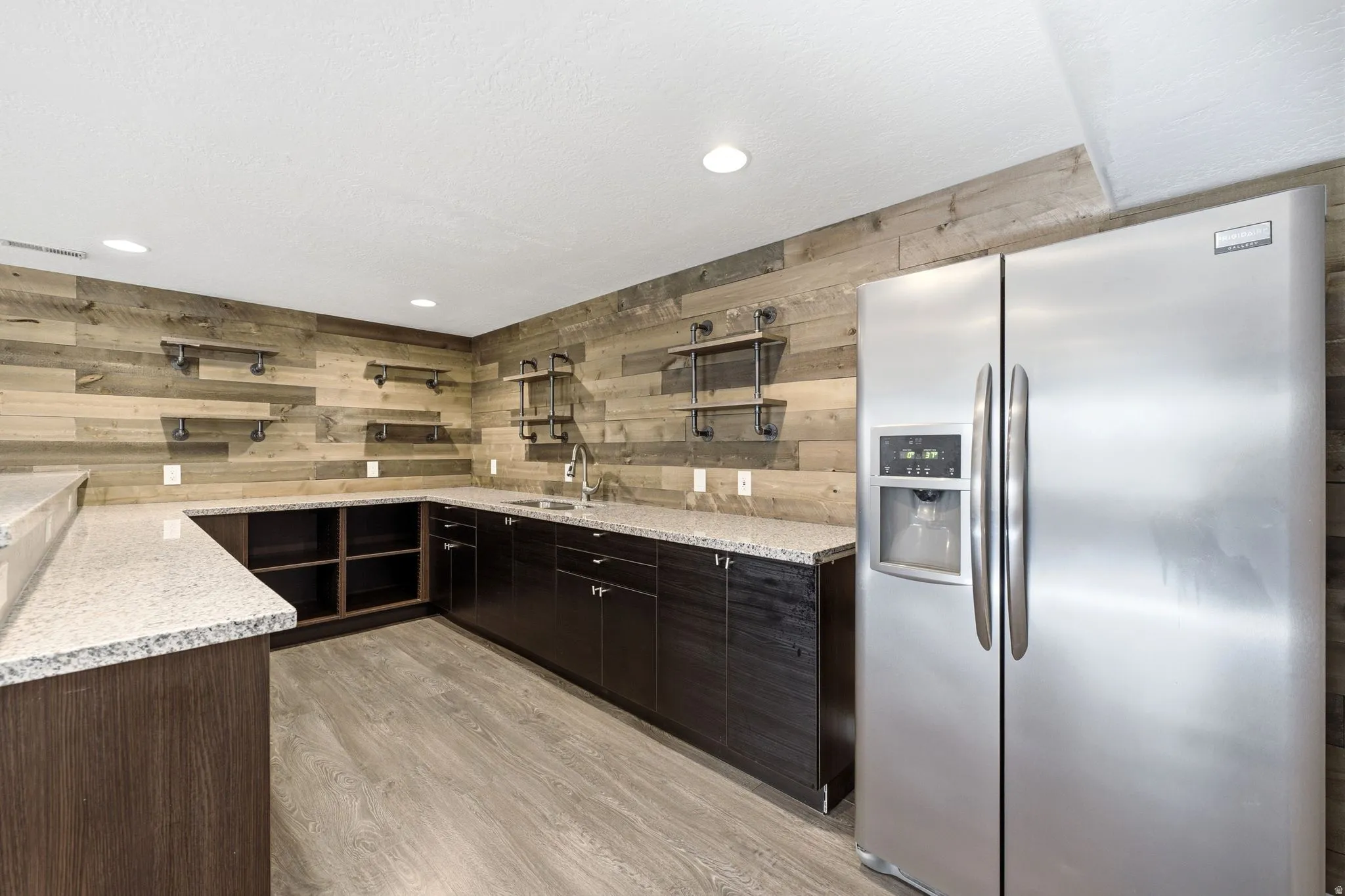 Wet bar with wood-finish flooring, L-shaped granite-style countertops, dark wood-finish cabinetry, a stainless steel sink, and a stainless steel side-by-side refrigerator