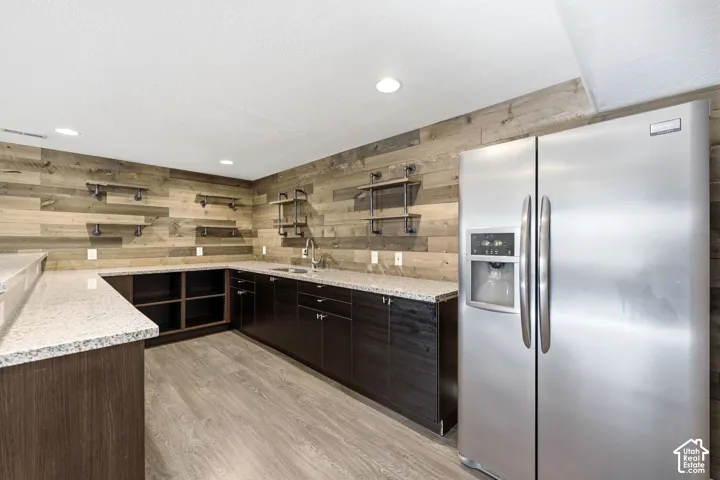 Wet bar with wood-finish flooring, L-shaped granite-style countertops, dark wood-finish cabinetry, a stainless steel sink, and a stainless steel side-by-side refrigerator