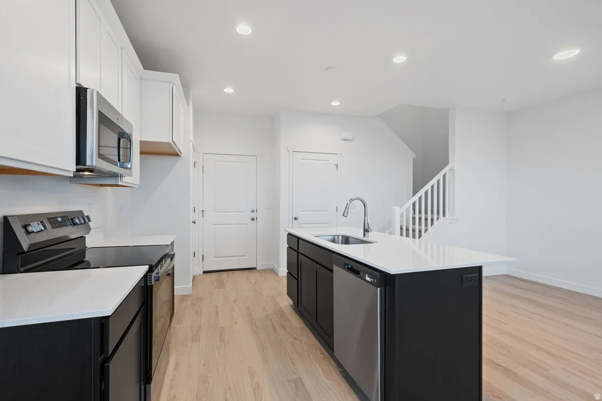 Contemporary kitchen featuring light wood-finish flooring, white cabinetry, dark lower cabinetry, stainless steel appliances, and an island with an integrated sink