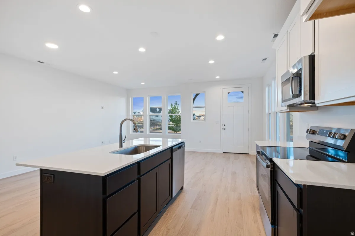Open-concept kitchen featuring a dark-toned island with an undermount sink, light countertops, stainless steel appliances, and light wood-finish flooring