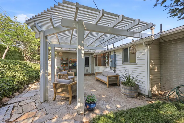 Expansive flagstone patio beneath a louvered pergola, featuring white siding, a brick exterior wall, and surrounding mature landscaping