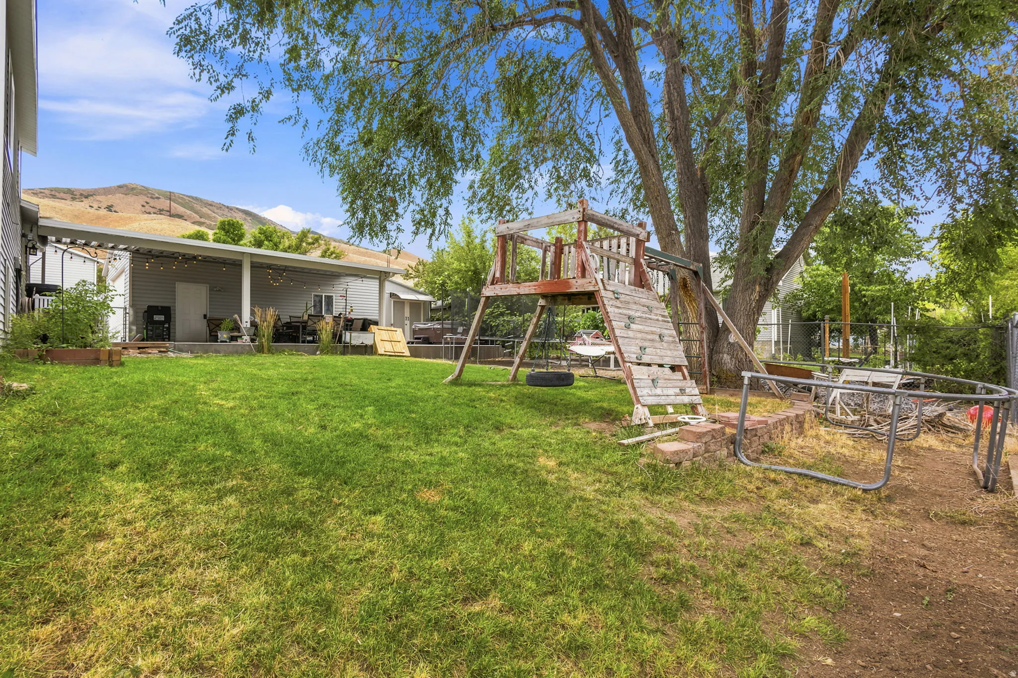 Expansive lawn area with a large mature tree, featuring a covered patio with string lighting, and raised garden beds