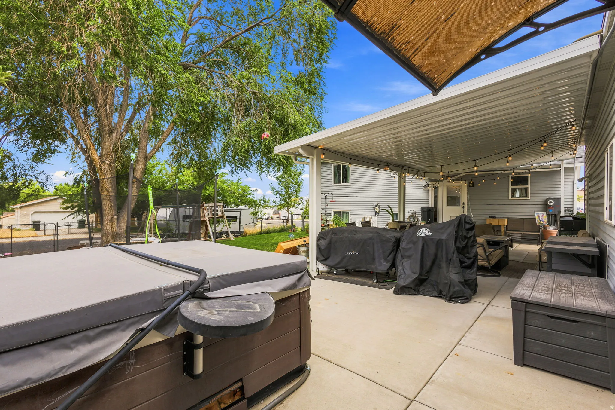 Spacious concrete patio featuring a covered seating area with string lighting, an uncovered area with a hot tub, and mature trees providing natural shade