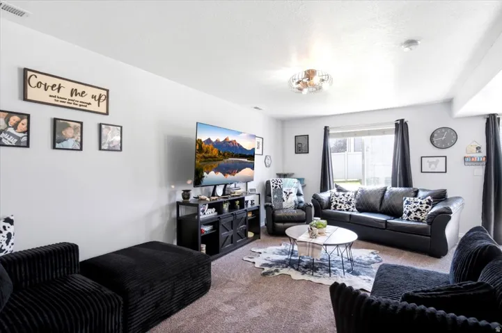 Spacious living area featuring light gray walls, plush carpet flooring, a multi-pane window with dark drapery, a built-in entertainment console, and a contemporary ceiling light fixture