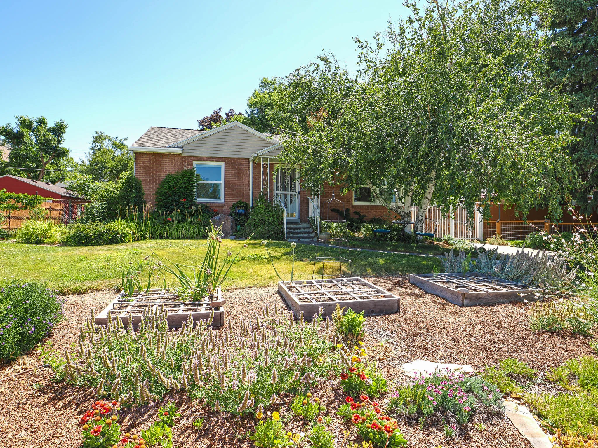 Front yard featuring multiple raised garden beds with established plantings, mature trees, and a brick-faced residence with a gabled roofline