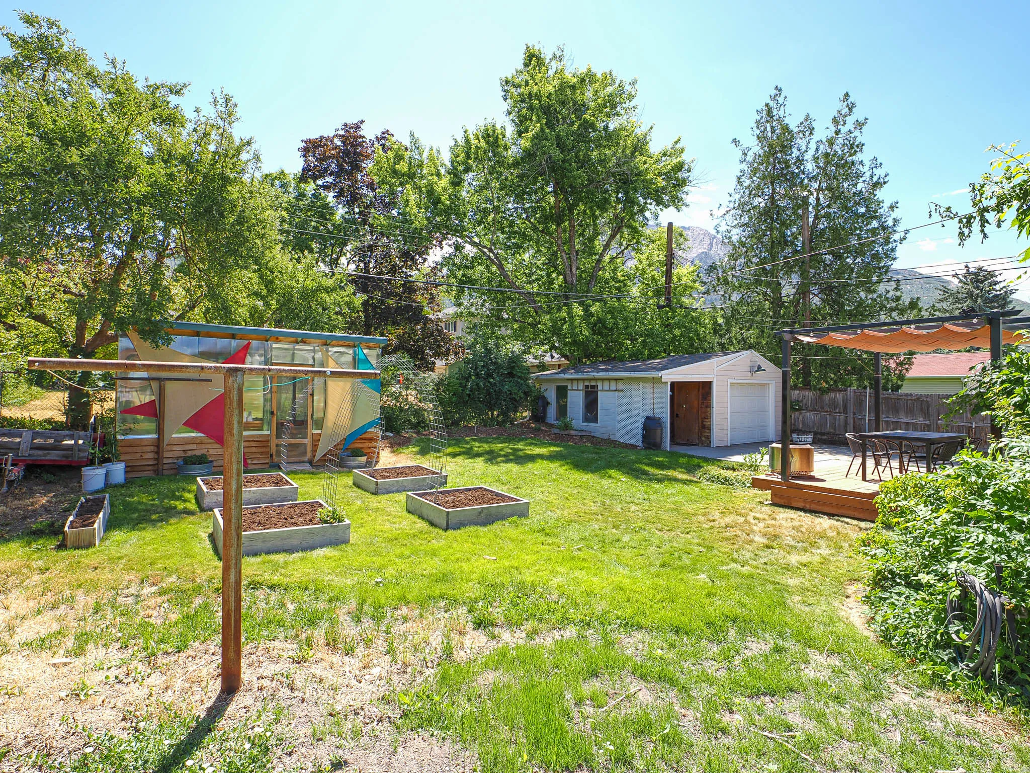 Spacious backyard featuring a detached garage, a greenhouse structure, multiple raised garden beds, a wood deck with a pergola, and mature trees