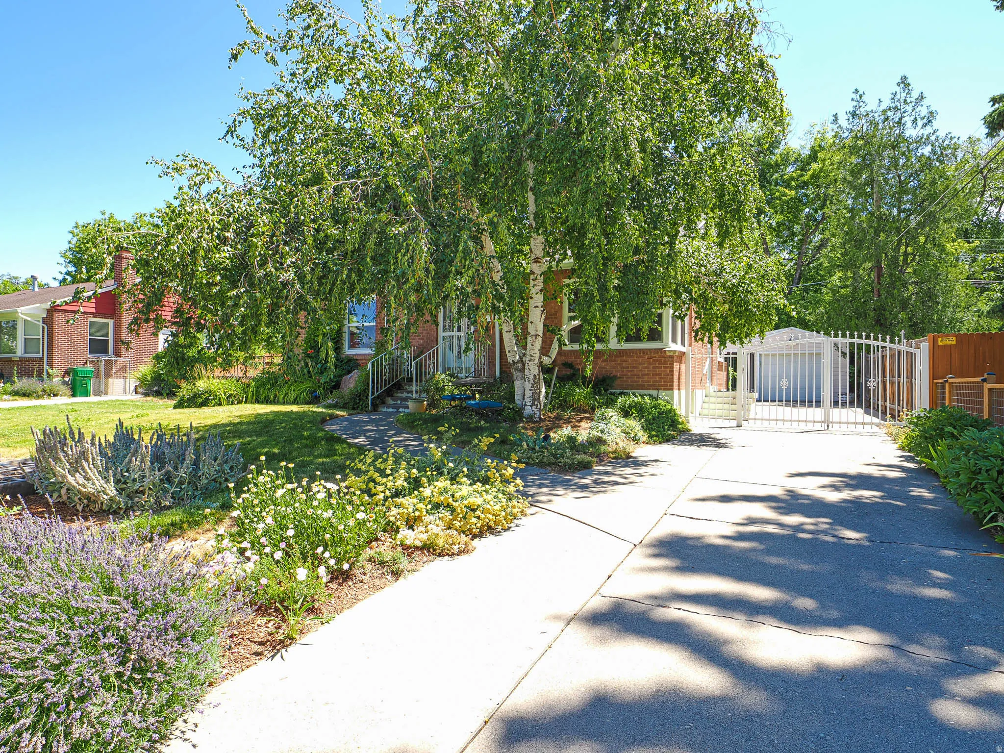Brick exterior residence with mature birch tree, landscaped front yard, paved driveway, and white metal privacy gate