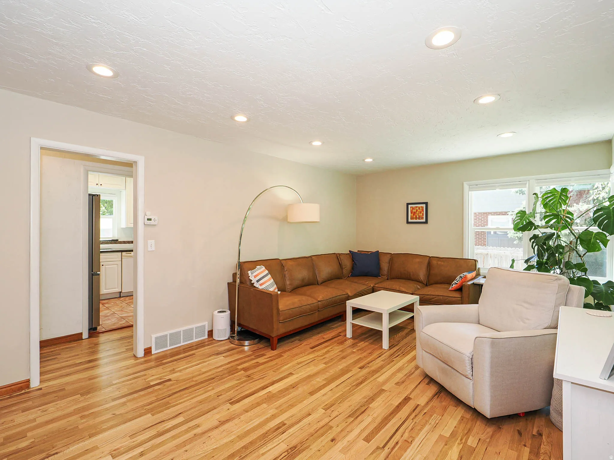 Spacious living area featuring wood-finish flooring, recessed ceiling lighting, ample natural light from a large window, and a clear entryway to an adjacent kitchen