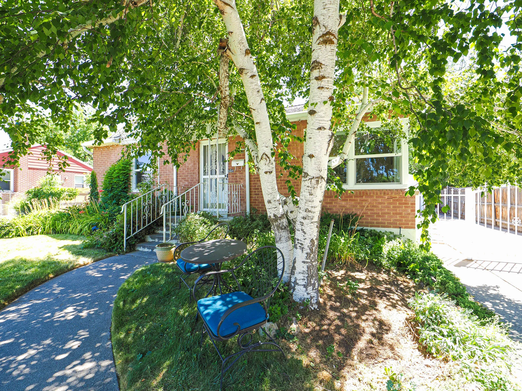 Brick exterior featuring mature shade trees, a paved walkway, front entry steps with metal railings, and a security screen door