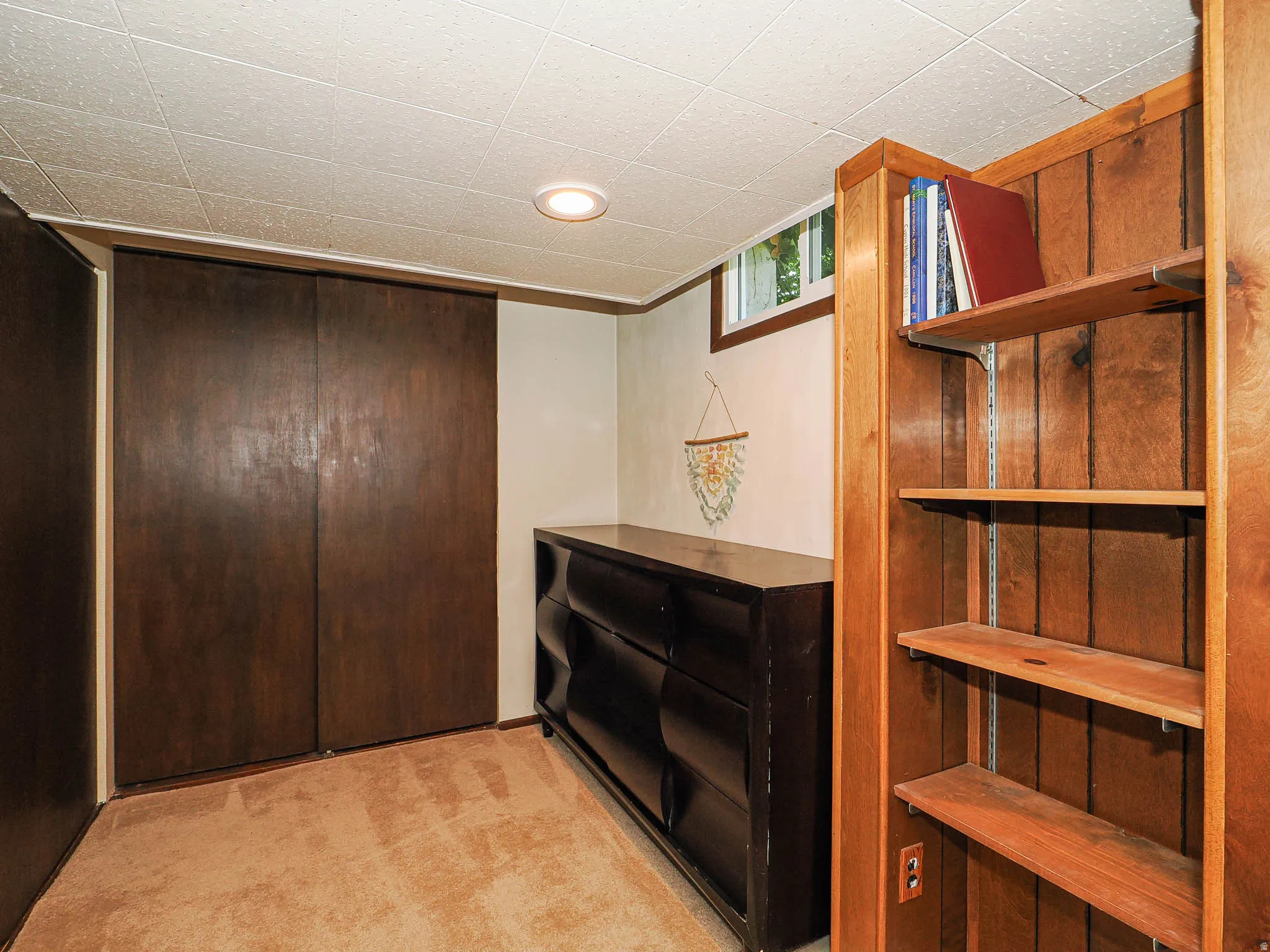 Recessed ceiling lighting illuminates a room featuring a drop ceiling, brown bi-fold closet doors, wall-mounted shelving, and wood paneling