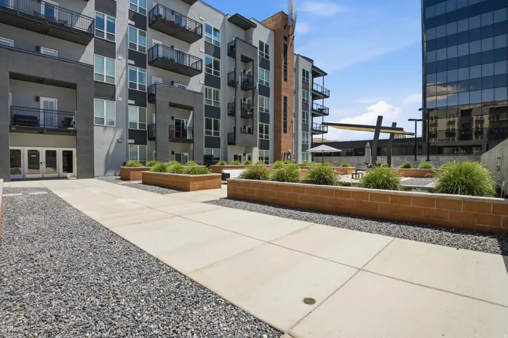 Expansive paved patio area featuring a modern pergola structure, brick planters with greenery, and gravel accents, set against a contemporary multi-story building