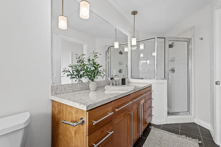 Contemporary bathroom featuring a wood-finish vanity with a light granite countertop, dual pendant lighting, and a glass-enclosed shower with white tiled walls