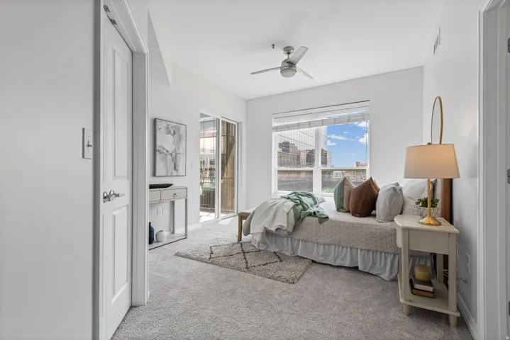 Carpeted interior space featuring a large window with blinds, a sliding glass door to an exterior balcony, a ceiling fan, and white paneled doors