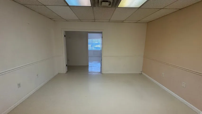 Spacious interior room featuring light-colored flooring, neutral wall tones, drop ceiling with fluorescent lighting, and an open doorway to an additional interior space