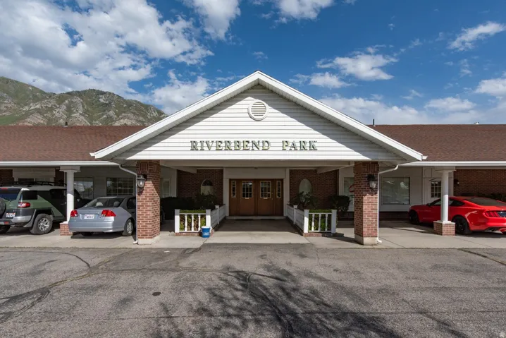 Building exterior featuring a gabled entrance with "RIVERBEND PARK" signage, brick pillars, wood-finish double entry doors, and white siding