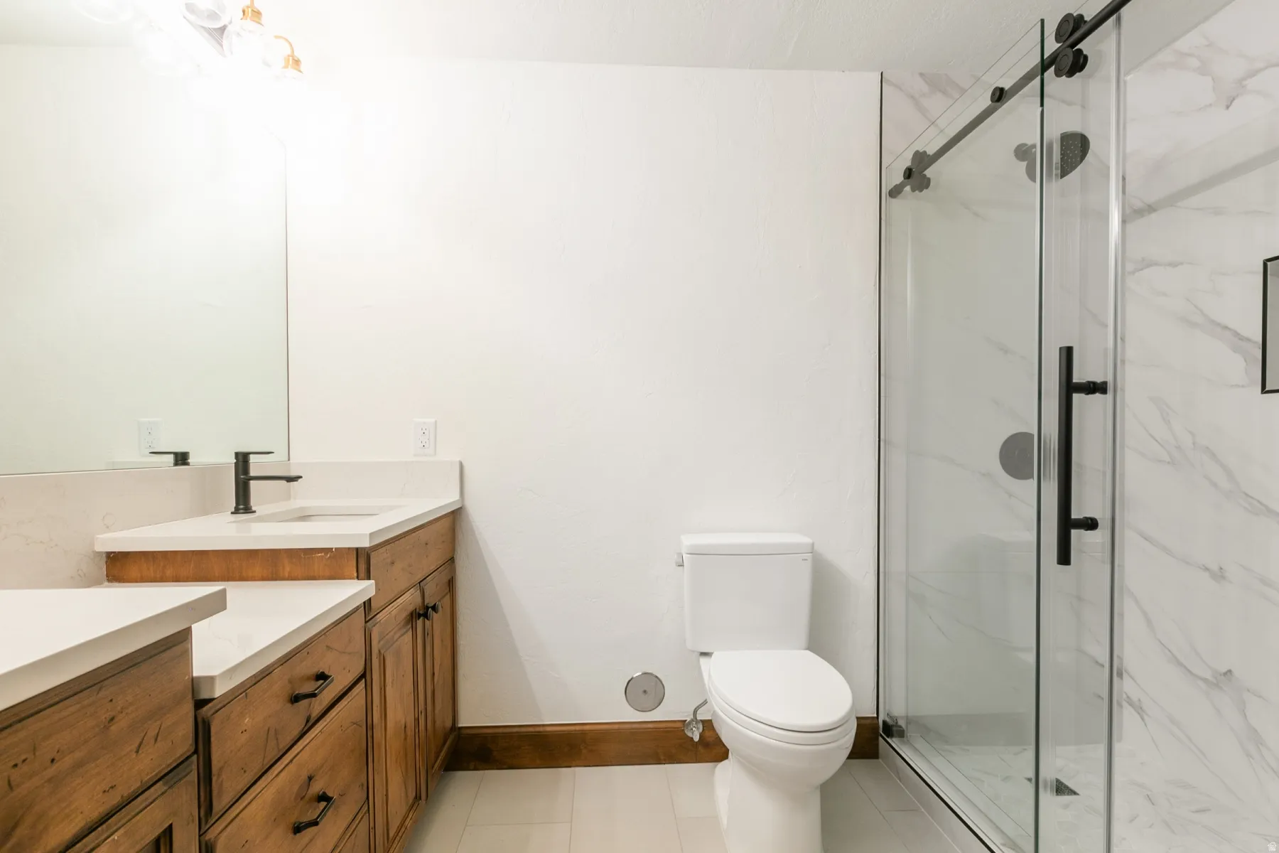 Bathroom featuring a dual vanity with light-toned countertops and wood cabinetry, a frameless glass shower enclosure with matte black hardware, and large format wall tiling