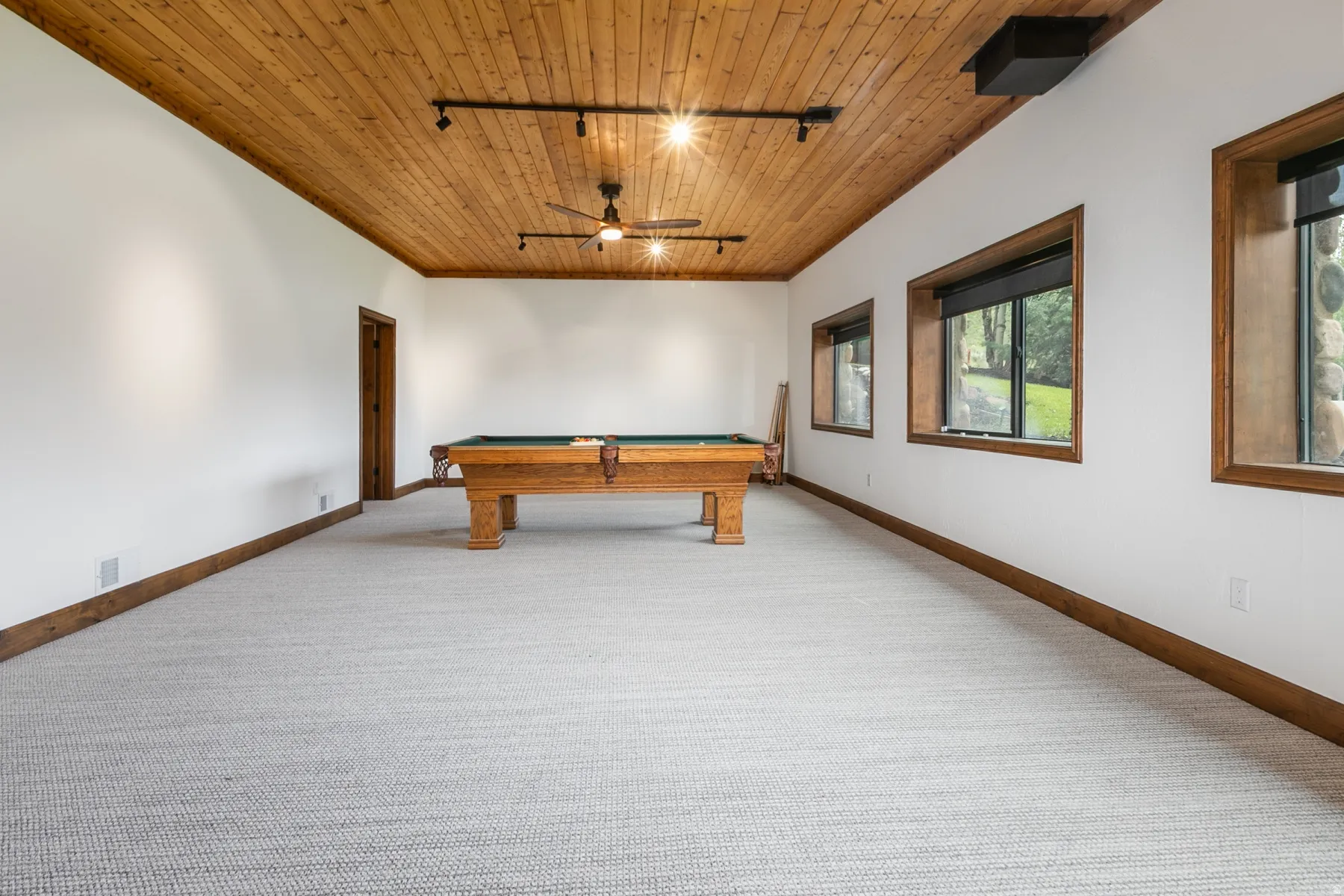 Spacious recreational room featuring a wood plank ceiling with track lighting, recessed windows with wood trim, and light-toned carpet flooring