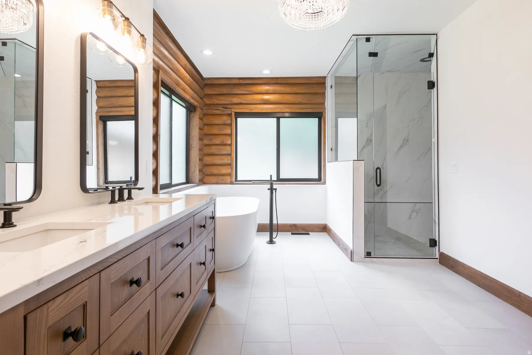 Spacious bathroom featuring a dual vanity with a light-toned countertop and wood-finish cabinetry, a freestanding soaking tub, and a glass-enclosed shower with tiled walls