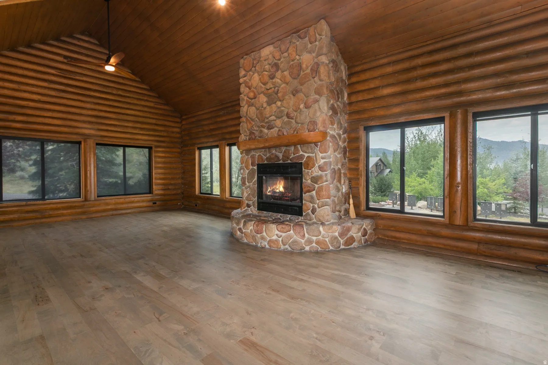 Expansive living area with a stone fireplace, wood-finish flooring, log walls, a vaulted wood-paneled ceiling, and multiple window installations