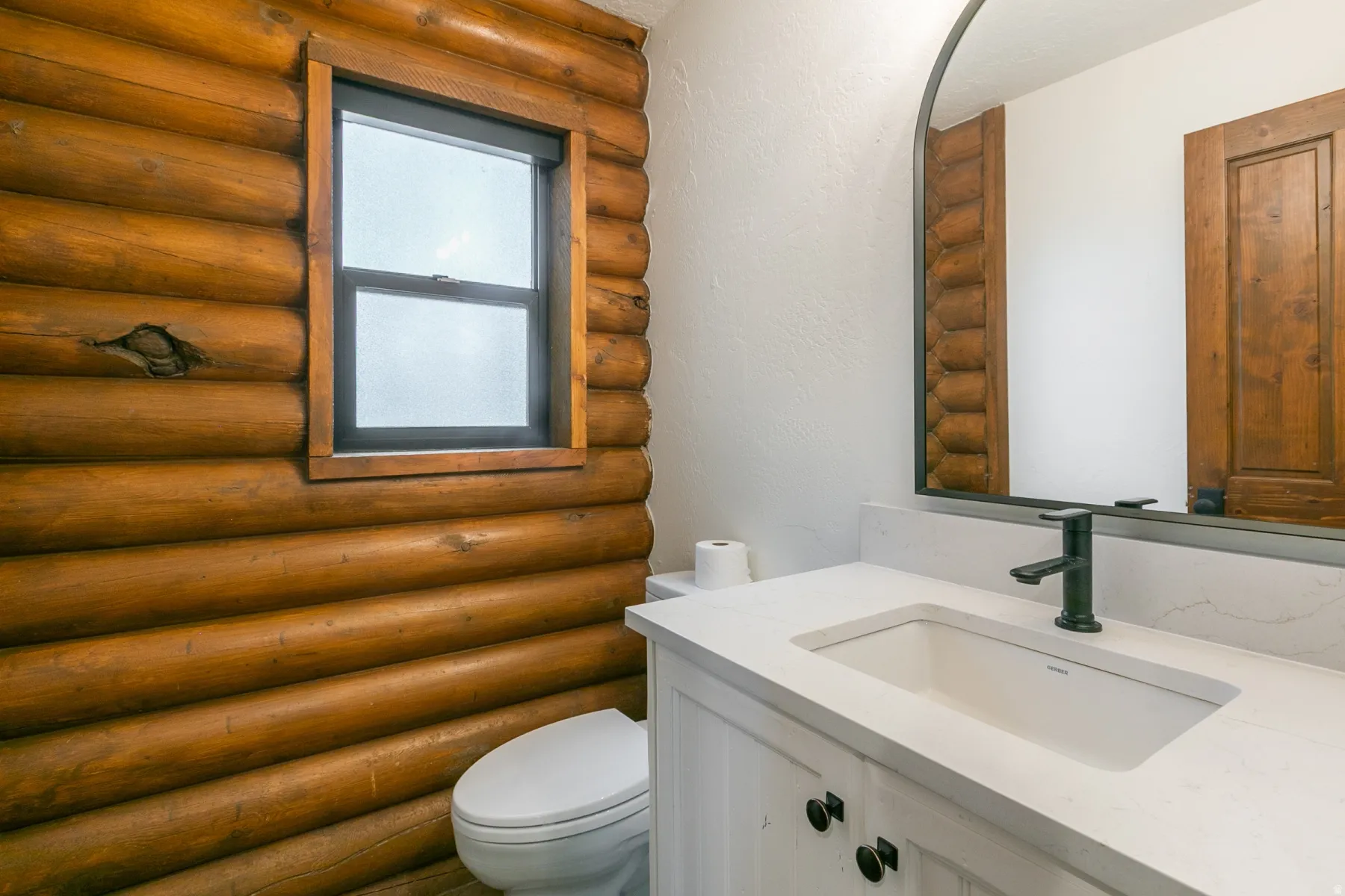 Bathroom featuring log wall construction, a white vanity with panel doors and dark hardware, an integrated sink with a matte black faucet, a light-toned countertop, and an arched mirror