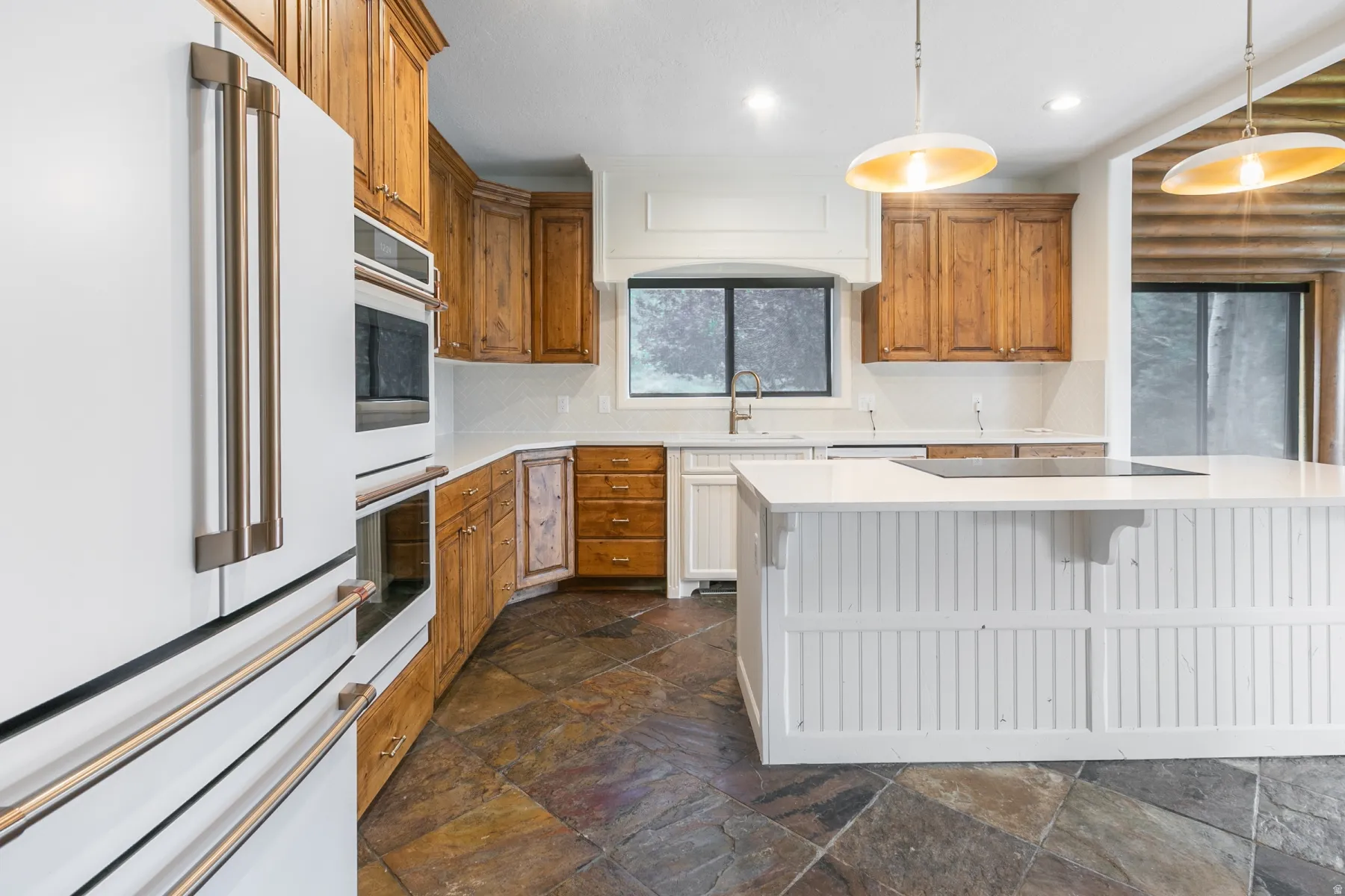 Kitchen featuring a white refrigerator with brushed metal hardware, built-in double ovens, natural wood cabinetry, a white countertop island with integrated cooktop, and slate-finish tile flooring