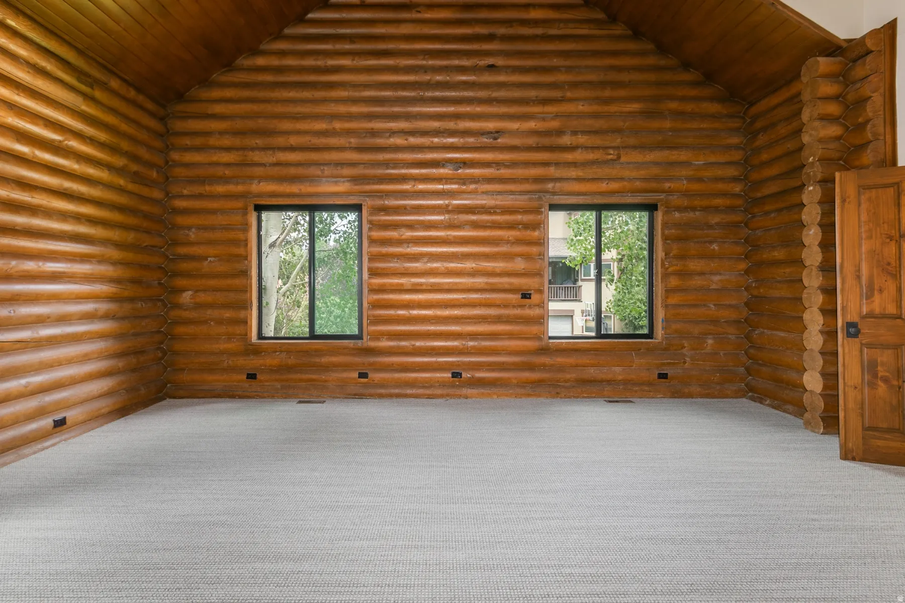 Room featuring log wall construction, a vaulted log ceiling, two windows with dark frames, and light-colored carpet flooring