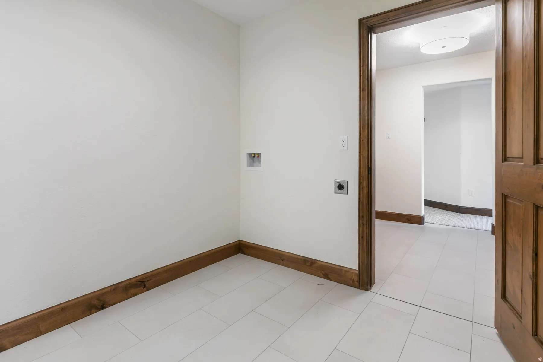 Utility room featuring light-toned rectangular floor tiles, crisp white walls, and rich wood-finish baseboards