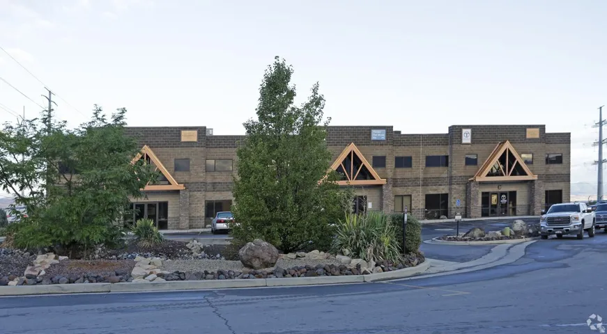 Exterior brick facade building featuring gabled architectural accents with exposed wood beams, a paved parking area, and landscaped rock gardens with mature trees