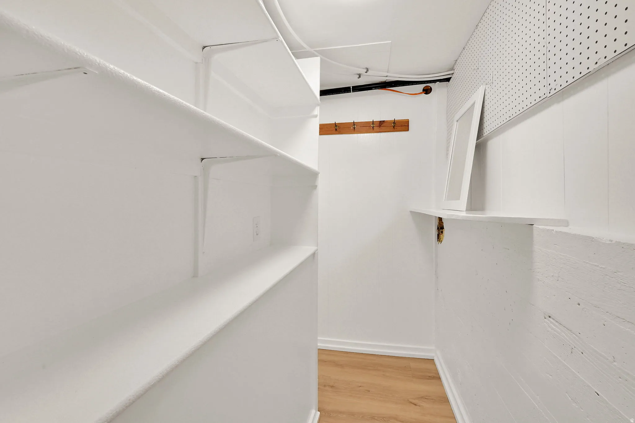 Dedicated storage area featuring extensive built-in shelving, durable wood-finish flooring, a wall-mounted coat rack, a wall-mounted shelf, and pegboard paneling