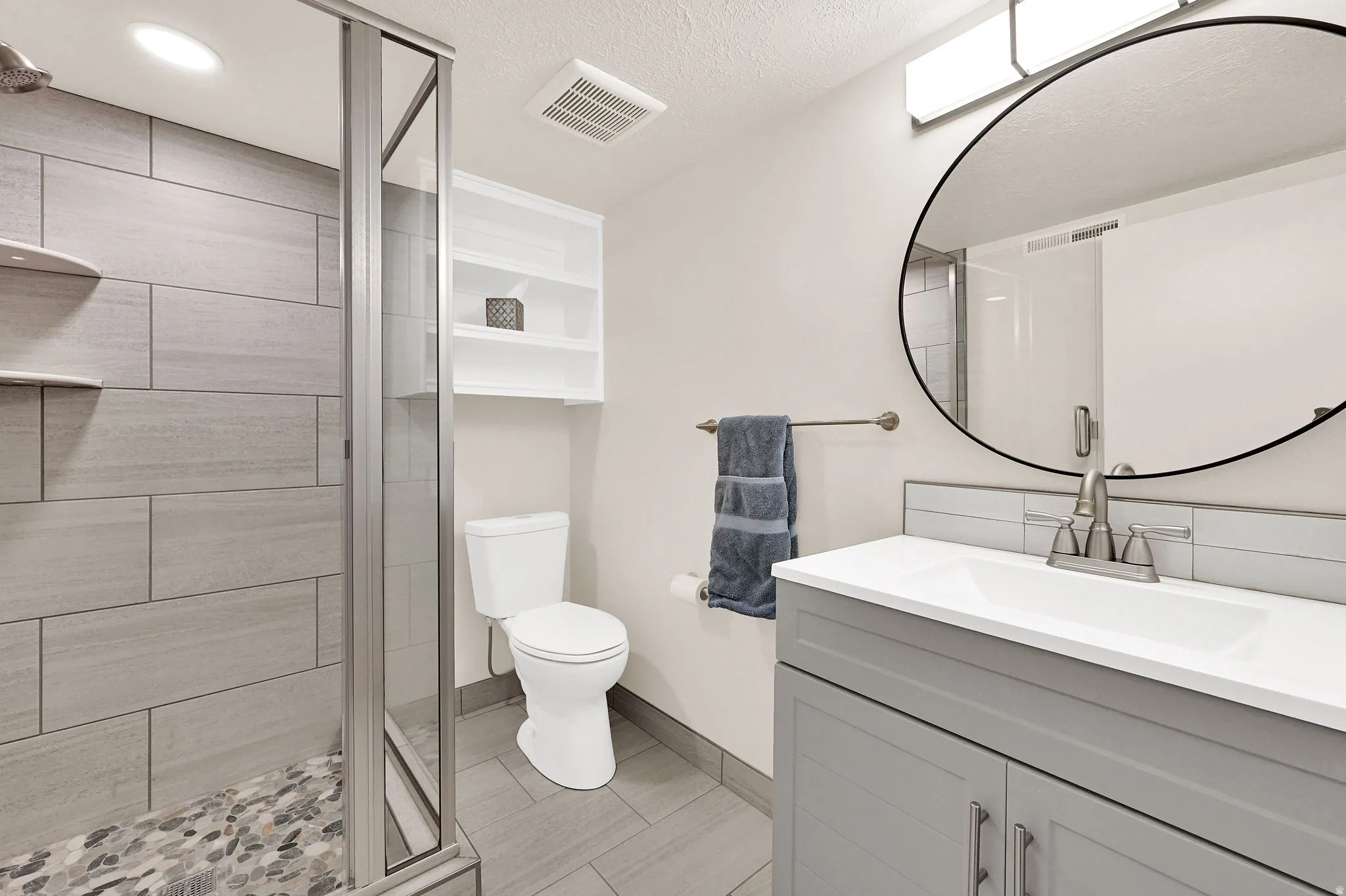 Contemporary bathroom featuring a glass-enclosed shower with large format tile surround and pebble floor, a single vanity with a white countertop and grey cabinetry, a circular wall mirror, and a wall-mounted shelving unit