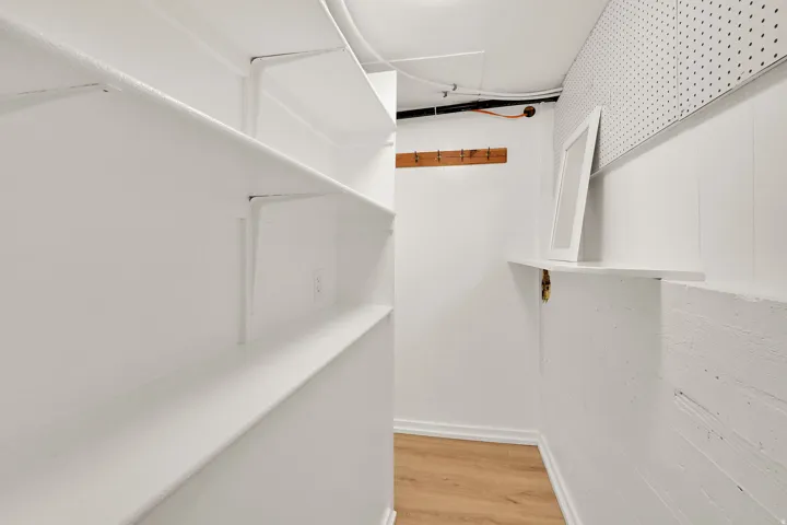 Dedicated storage area featuring extensive built-in shelving, durable wood-finish flooring, a wall-mounted coat rack, a wall-mounted shelf, and pegboard paneling