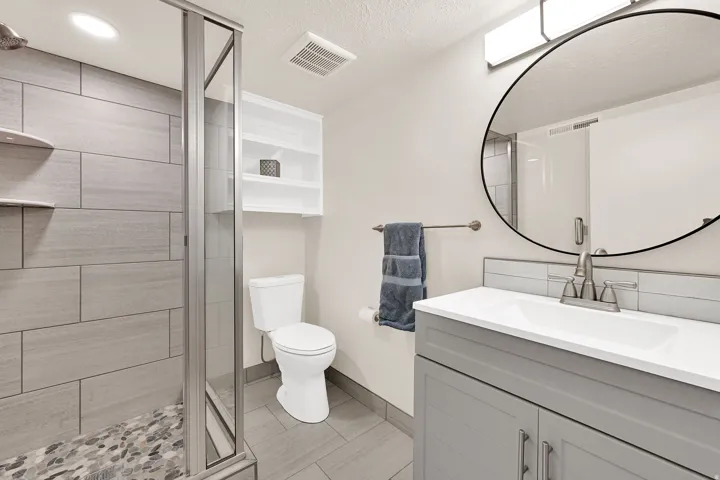 Contemporary bathroom featuring a glass-enclosed shower with large format tile surround and pebble floor, a single vanity with a white countertop and grey cabinetry, a circular wall mirror, and a wall-mounted shelving unit