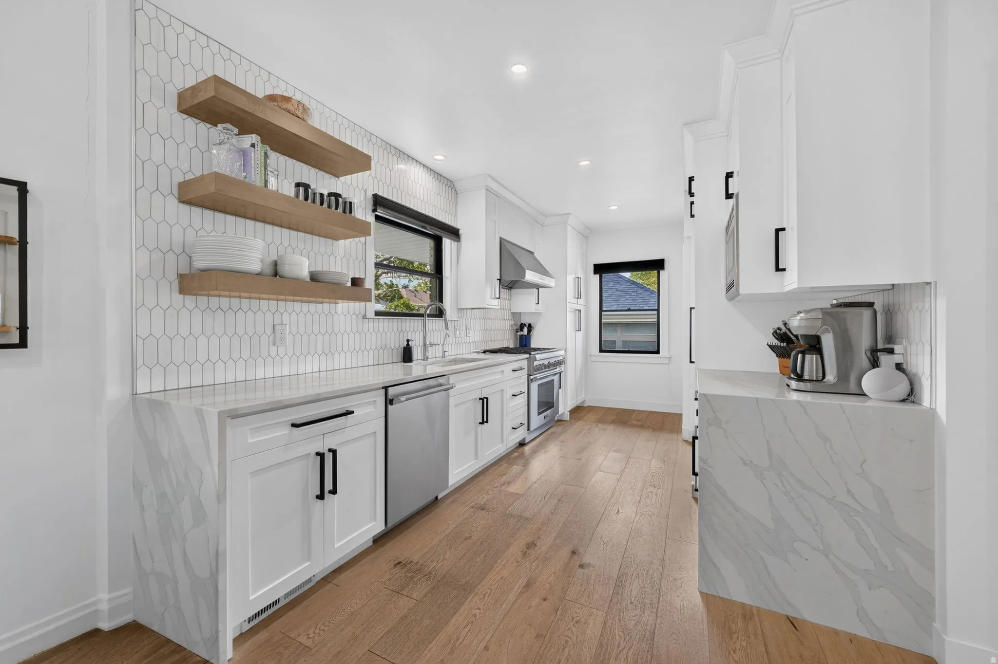 Stunning modern kitchen featuring white cabinetry, counter to ceiling tile backsplash, wood accent floating shelves, stainless steel appliances, and waterfall quartz countertops.